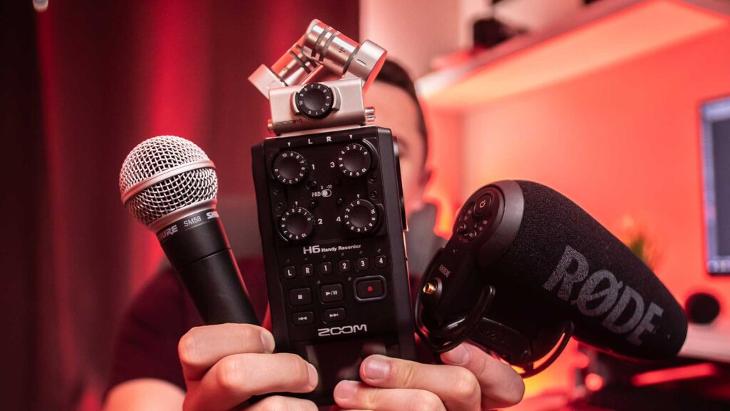 Close-up of a person holding a Zoom H6 Handy Recorder, a Shure microphone, and a RØDE microphone for professional audio recording.